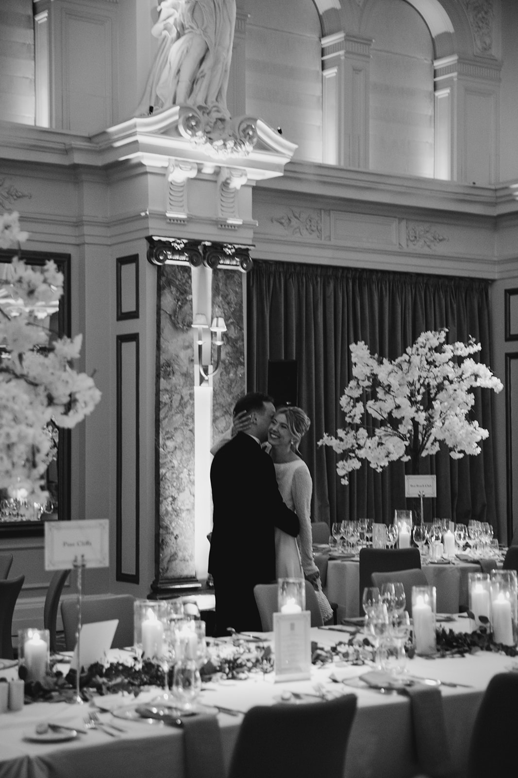 Bride and groom embracing in reception space with bride wearing elegant updo and soft luminous makeup among candlelit tables