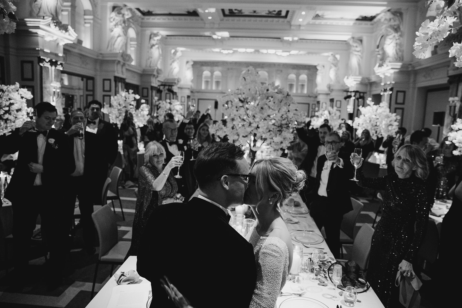 Bride and groom kissing at reception table surrounded by guests toasting in grand London ballroom with bride wearing relaxed updo and glowing makeup