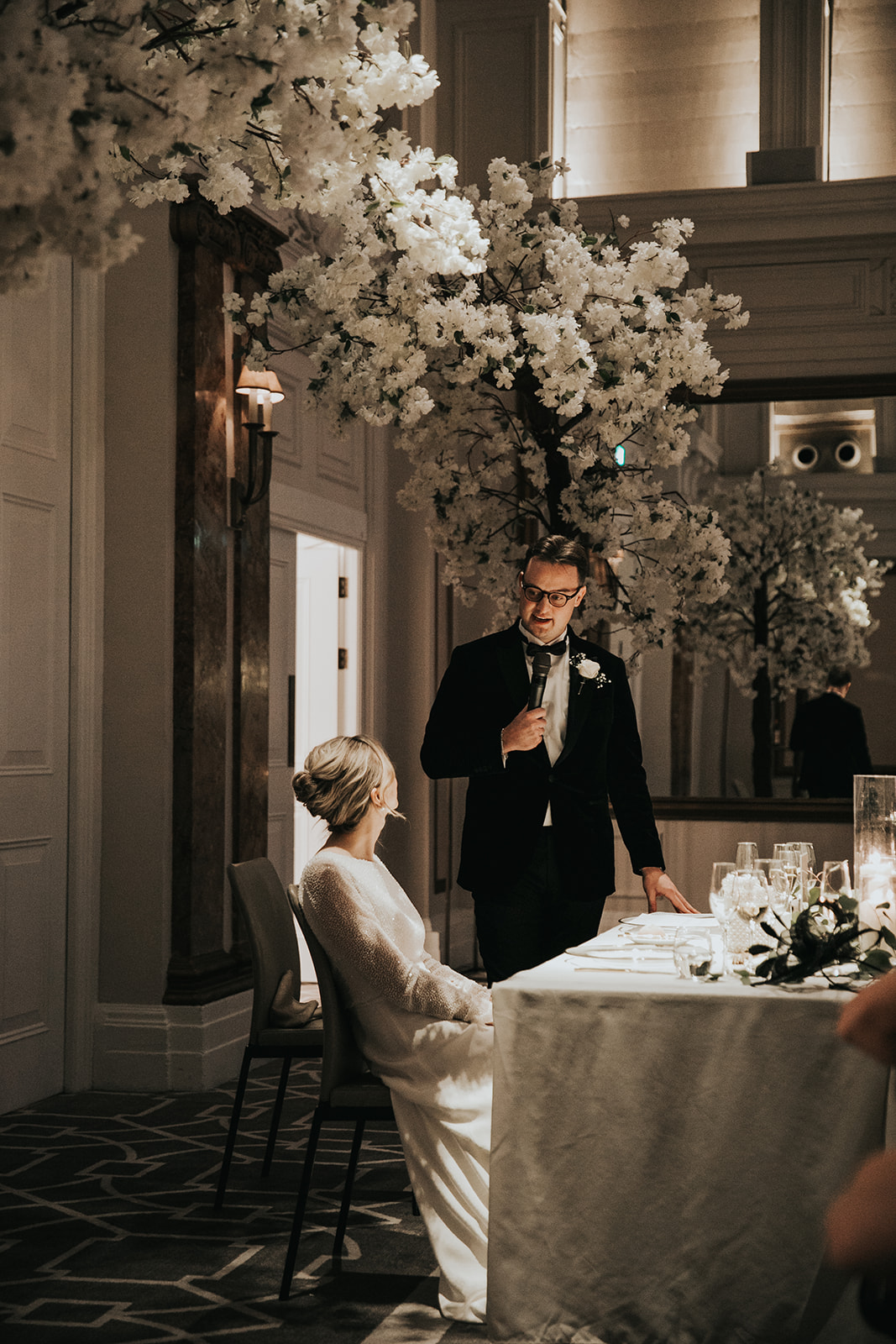 Groom giving wedding speech with bride seated beneath floral installation at Kimpton Fitzroy London with elegant updo and soft natural makeup