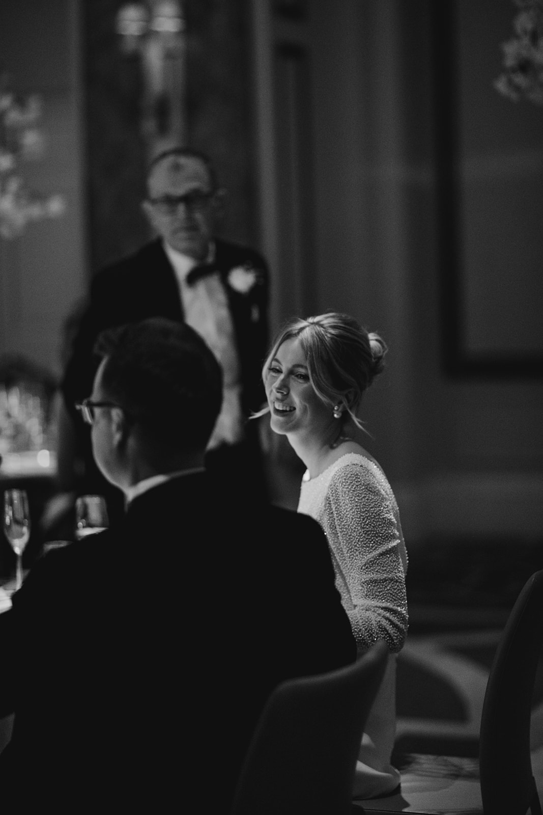 Bride smiling during speeches in black and white moment at London wedding with soft bridal makeup and loosely styled updo
