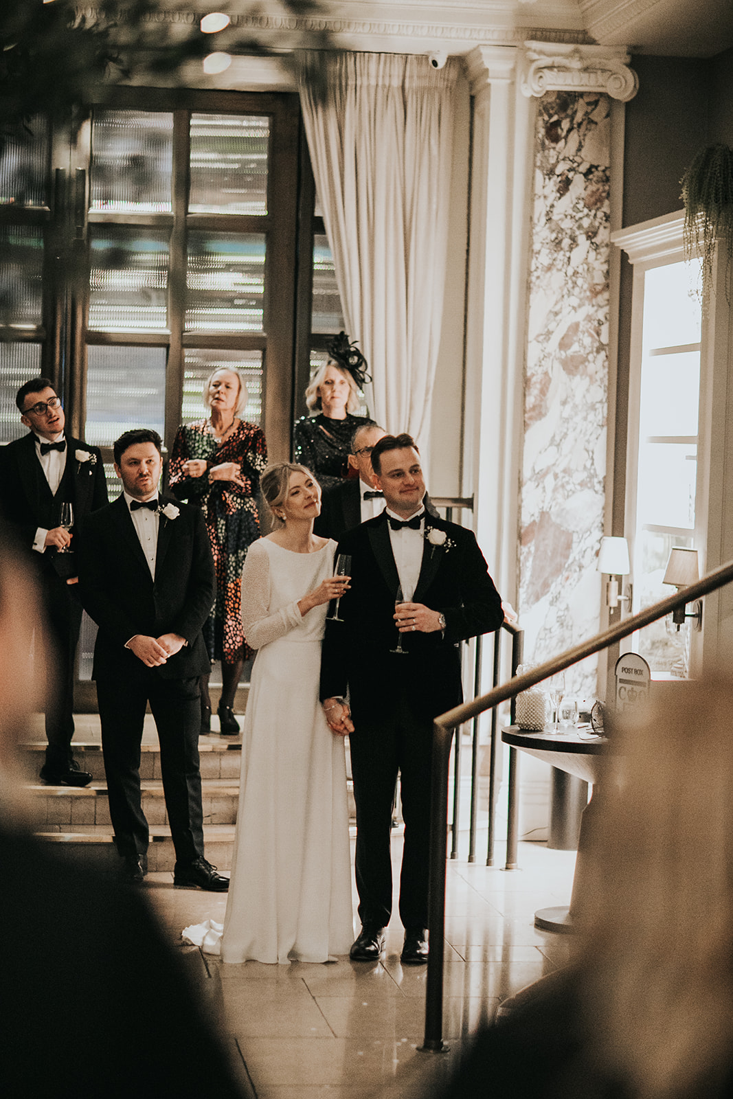 Bride and groom listening to speeches together on staircase at Kimpton Fitzroy London with bride wearing classic updo and radiant natural makeup