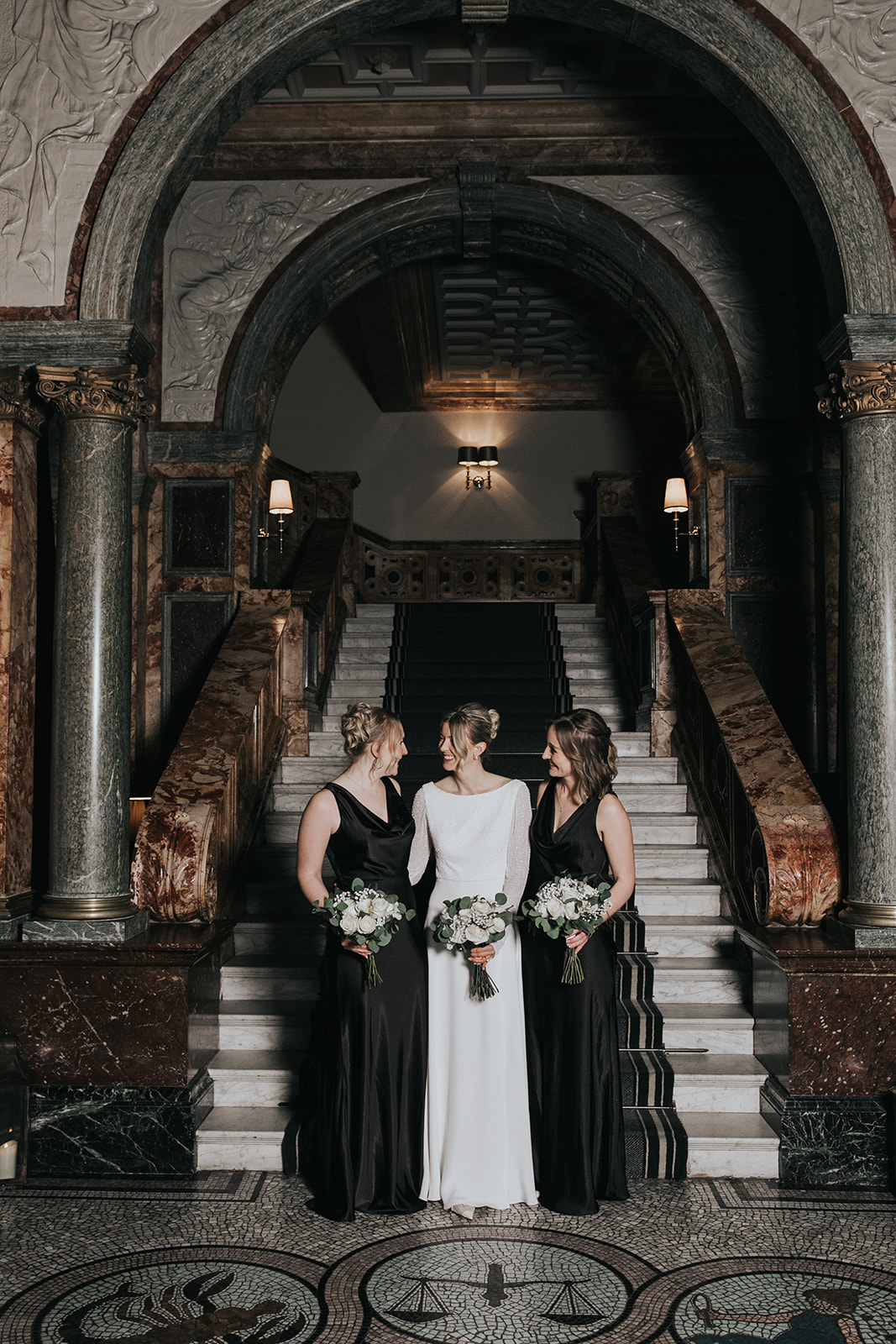 Bridal party portraits on staircase at Kimpton Fitzroy London with bride wearing elegant updo and natural glowing makeup alongside bridesmaids in black dresses