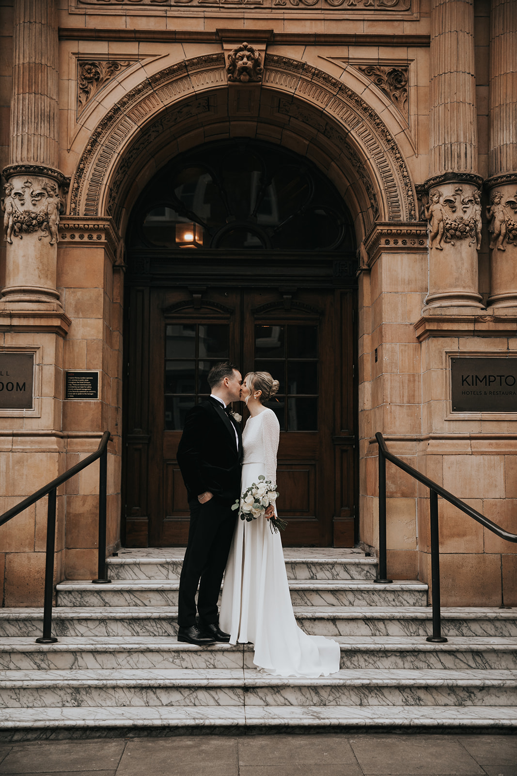 Bride and groom kissing on steps outside Kimpton Fitzroy London with bride wearing classic French twist and soft glowing makeup